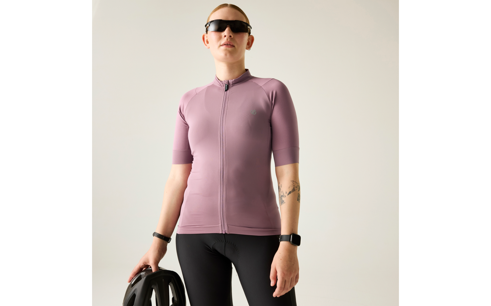 Person mit kurzen Haaren, trägt mauvefarbenes kurzärmeliges Reißverschluss-Radtrikot, schwarze Fahrradhose und Sonnenbrille, hält einen schwarzen Fahrradhelm, Tätowierung am Unterarm sichtbar, heller einfarbiger Hintergrund