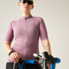 Person mit rosa-violettem Radtrikot und Sonnenbrille, hält Rennradlenker mit blauem Lenkerband; Tätowierung am Unterarm und schwarzes Armband/Smartwatch, heller Hintergrund