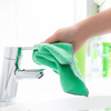 Hand hält grünes Mikrofasertuch an verchromten Wasserhahn über weißem Waschbecken, im Hintergrund unscharfe Flaschen