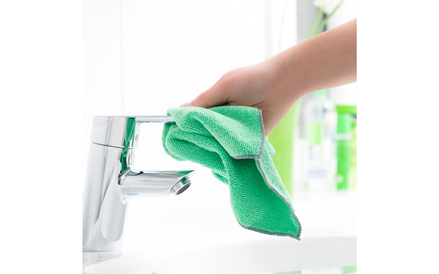 Hand hält grünes Mikrofasertuch an verchromten Wasserhahn über weißem Waschbecken, im Hintergrund unscharfe Flaschen