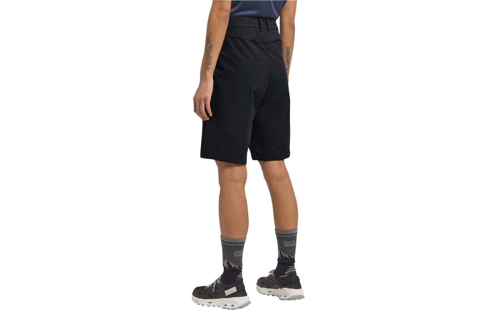 Rückansicht einer Person von der Taille abwärts, schwarze knielange Shorts, graue gemusterte Socken, schwarze Sneakers mit weißen Sohlen; sichtbare Unterarme mit Tätowierungen