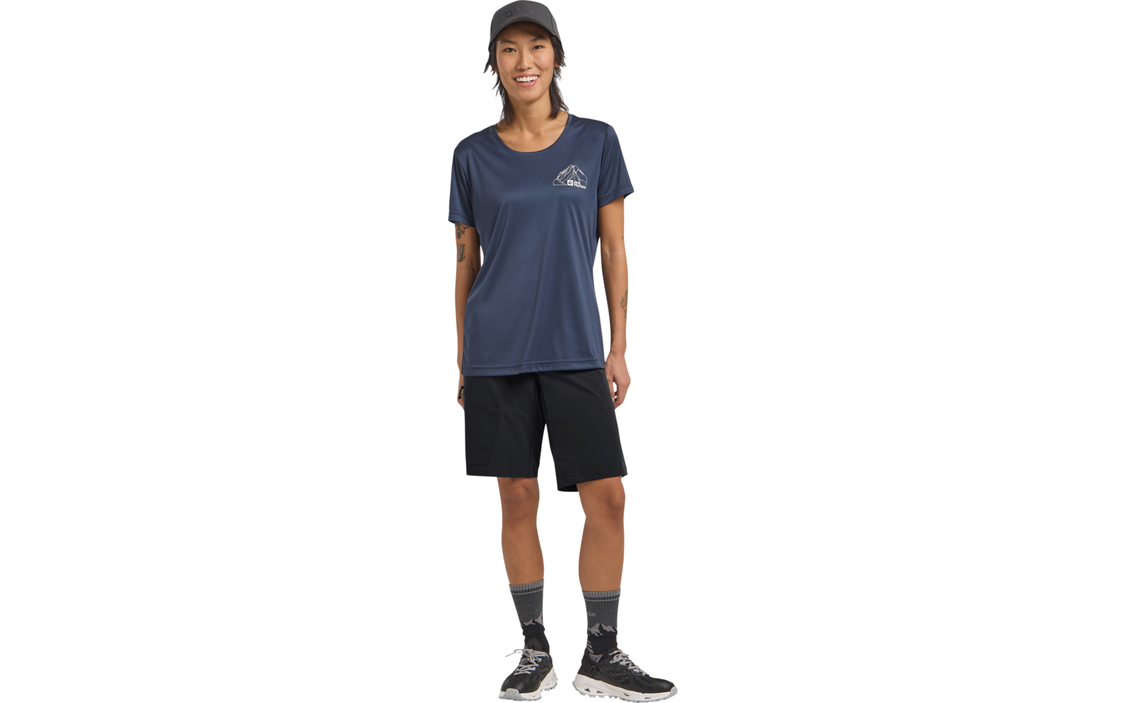 Person mit dunkler Cap, dunkelblauem T‑Shirt mit kleinem Brustlogo, schwarzen Shorts, grauen Socken und Sportschuhen, steht frontal