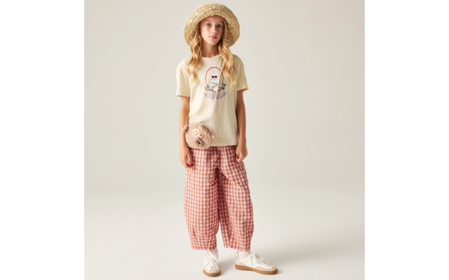 Stehendes blondes Mädchen vor hellem Hintergrund, trägt Strohhut, beiges T‑Shirt mit Grafik, rosa-weiß karierte weite Hose, weiße Sneaker und kleine runde Teddy-Umhängetasche