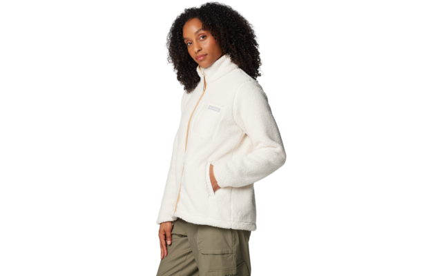 Person mit dunklem, lockigem Haar trägt weiße Fleecejacke mit Reißverschluss und Brusttasche, Hände in den Seitentaschen, olivgrüne Cargohose, Dreiviertelprofil vor hellem Hintergrund