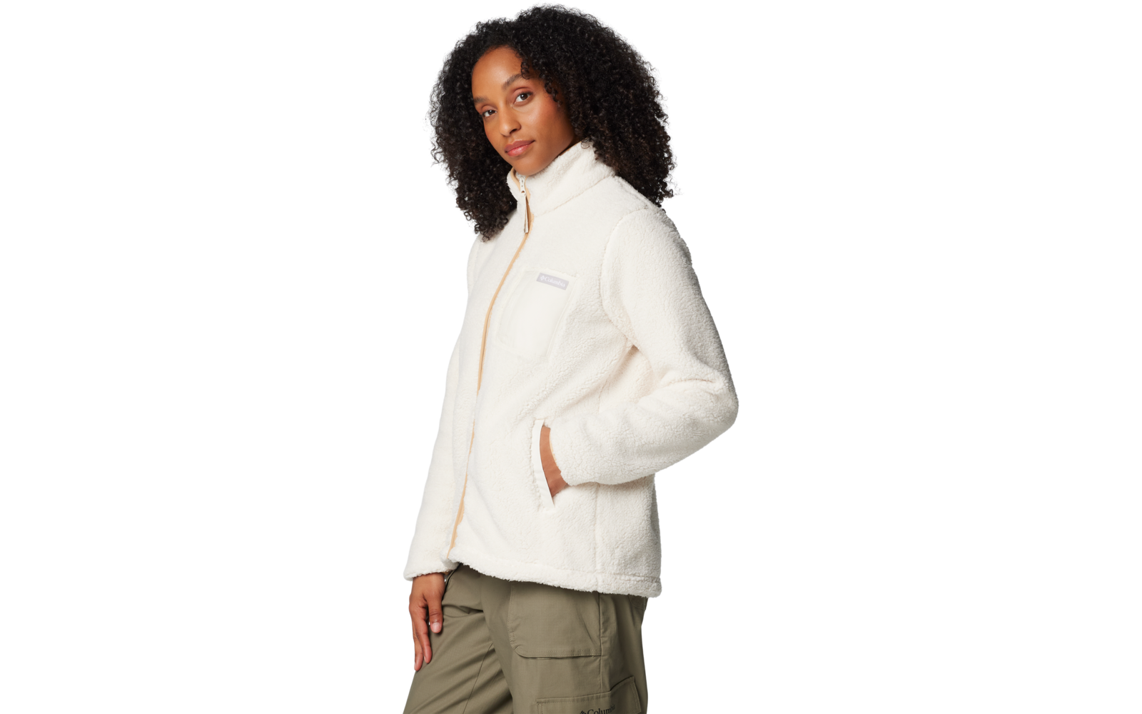 Person mit dunklem, lockigem Haar trägt weiße Fleecejacke mit Reißverschluss und Brusttasche, Hände in den Seitentaschen, olivgrüne Cargohose, Dreiviertelprofil vor hellem Hintergrund