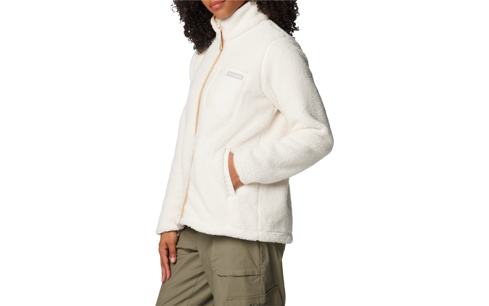 Personne aux cheveux bouclés foncés portant une veste polaire blanche avec fermeture éclair et poche poitrine, mains dans les poches latérales, pantalon cargo vert olive, trois-quarts profil sur fond clair