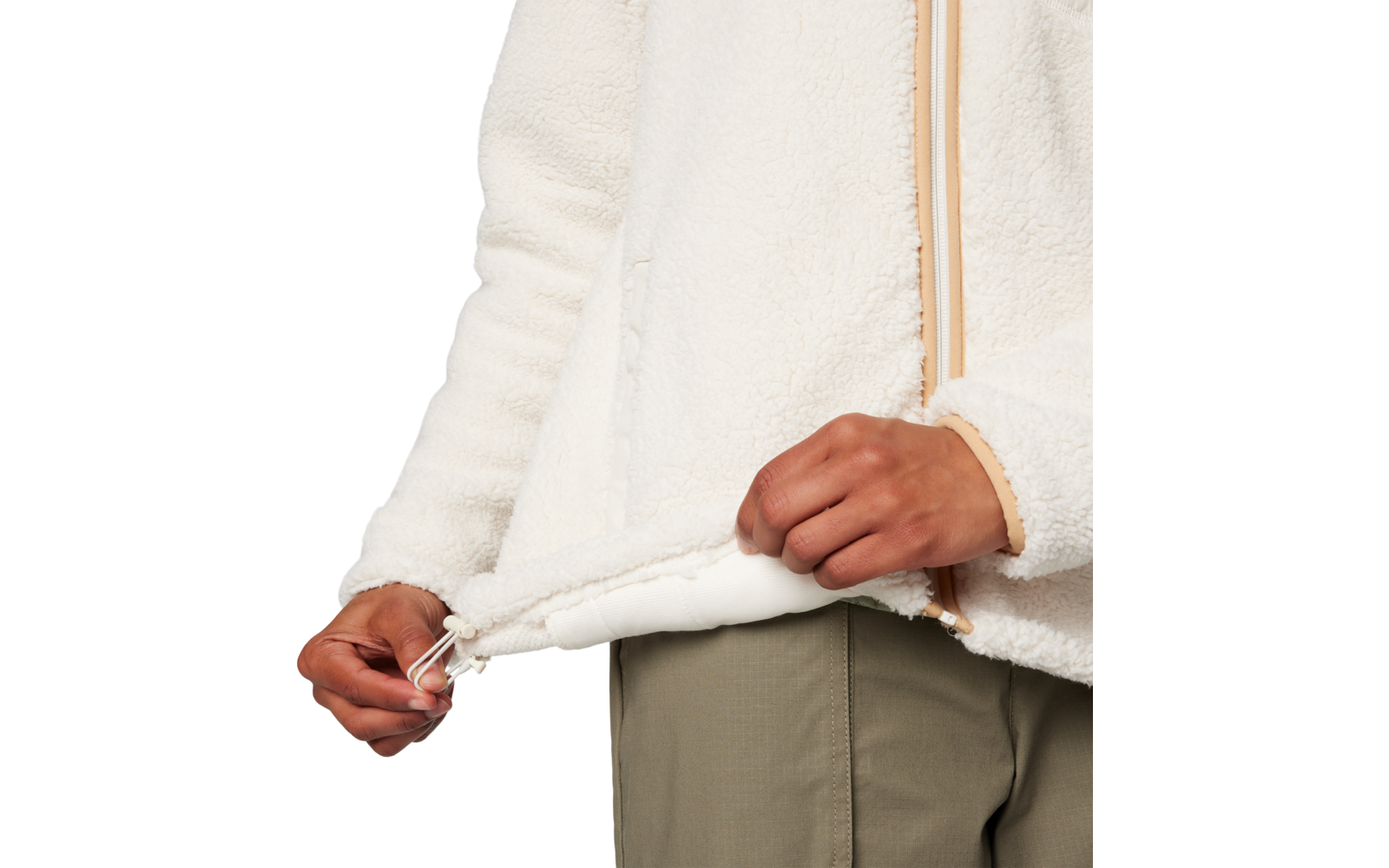 Oberkörper und Hände einer Person, die den Saum einer cremefarbenen Fleecejacke mit Reißverschluss und Kordelzug halten; olivgrüne Hose sichtbar