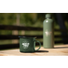 Tasse émaillée verte avec inscription 'camp life' et thermos vert sur une table en bois, clôture en bois floue et arbres en arrière-plan