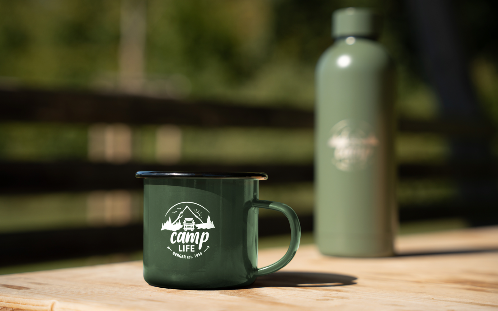 Grüner Emaille-Becher mit Aufdruck 'camp life' und grüne Thermosflasche auf einem Holztisch, verschwommener Holzzaun und Bäume im Hintergrund