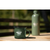 Grüner Emaille-Becher mit Aufdruck 'camp life' und grüne Thermosflasche auf einem Holztisch, verschwommener Holzzaun und Bäume im Hintergrund