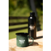 Grüner Emaille-Becher mit weißem "Camp Life"-Logo und schwarze Trinkflasche auf Holztisch, verschwommener grüner Hintergrund