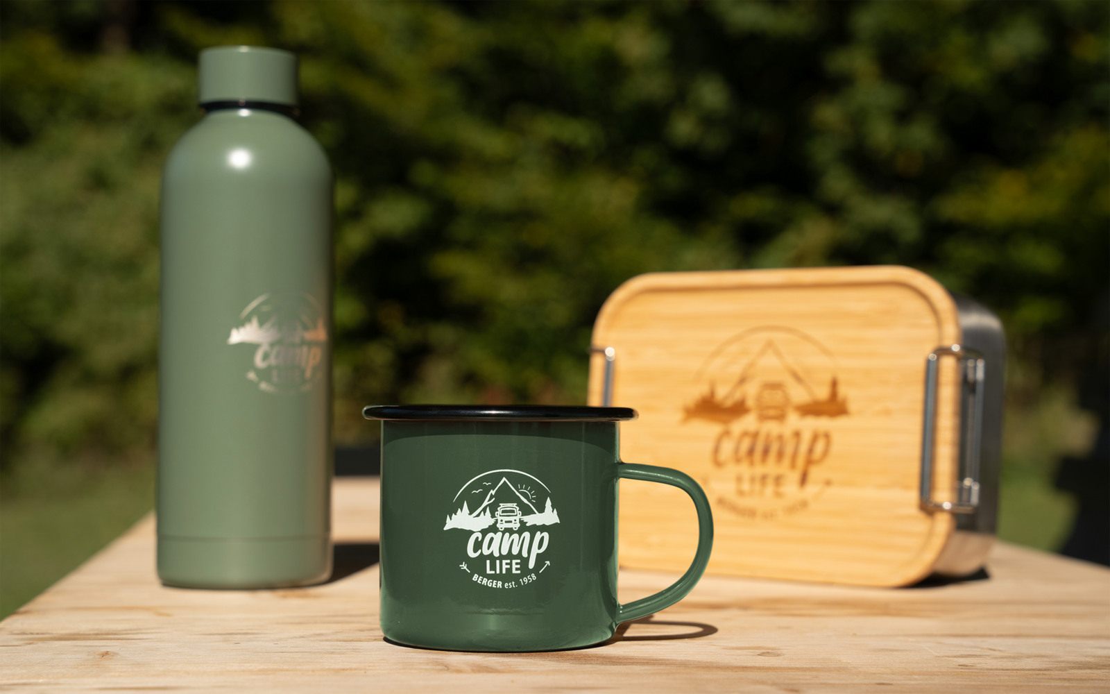 Grüne Trinkflasche, grüner Emaille-Becher mit Aufdruck „camp life“ und Holzbox mit Metallverschluss auf einem Holztisch, unscharfer grüner Hintergrund