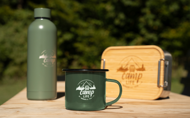 Grüne Trinkflasche, grüner Emaille-Becher mit Aufdruck „camp life“ und Holzbox mit Metallverschluss auf einem Holztisch, unscharfer grüner Hintergrund