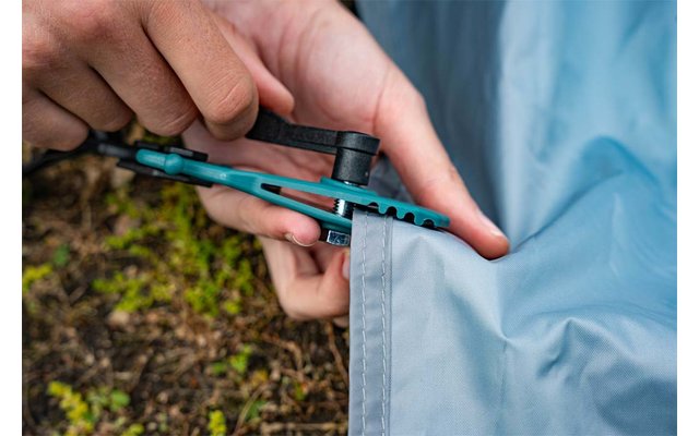 Zwei Hände halten ein blau-grünes Multitool an einer grau-blauen Zeltplane, das Werkzeug greift an einer Zeltöse; moosiger Erdgrund im Hintergrund