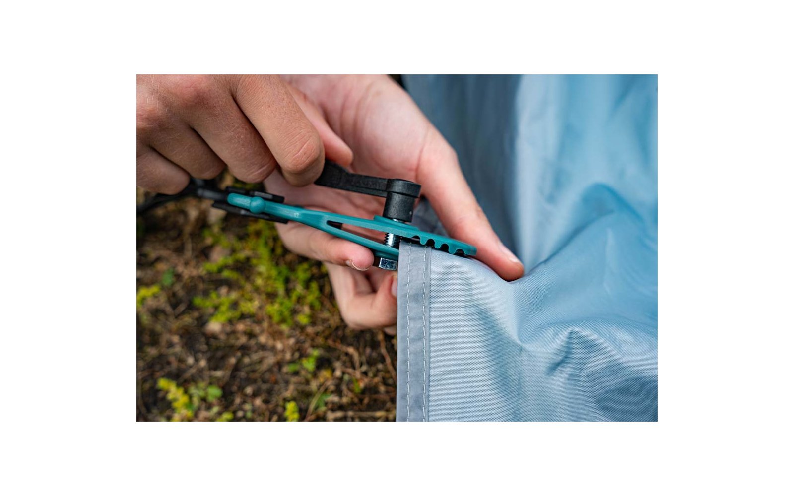 Zwei Hände halten ein blau-grünes Multitool an einer grau-blauen Zeltplane, das Werkzeug greift an einer Zeltöse; moosiger Erdgrund im Hintergrund