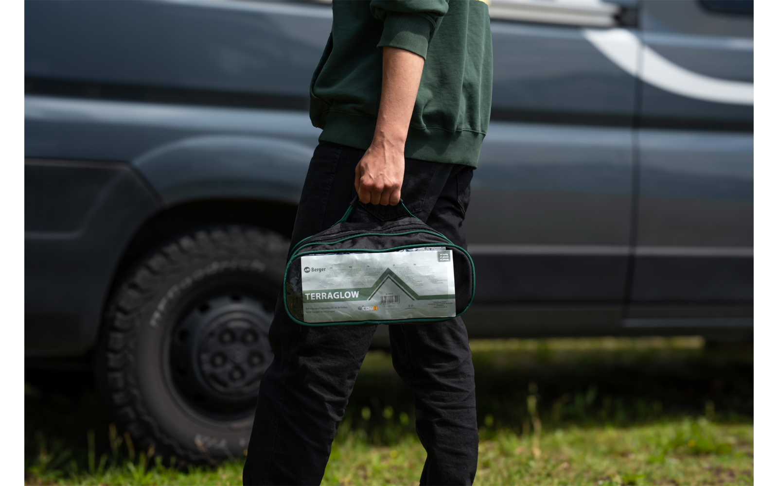 Teilweise sichtbare Person in grünem Oberteil und schwarzer Hose, hält eine durchsichtige Kulturtasche mit Aufdruck neben einem grauen Transporter