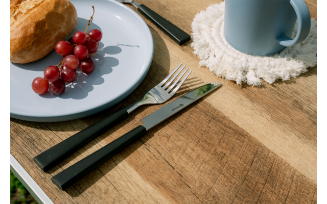 Blauw bord met rode druiven en een broodje, zwart mes en vork ernaast, blauwe mok op een witte onderzetter op een houten tafel