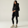 Donna in piedi su sfondo chiaro, indossa un gilet imbottito nero con cappuccio, top e leggings neri, stivaletti beige con lacci, mani davanti al corpo