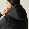 Persona di profilo con capelli scuri raccolti, indossa un gilet imbottito nero con cappuccio e una maglia a maniche lunghe scura, mano sul cappuccio, orecchino a cerchio dorato, sfondo chiaro neutro