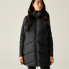 Persona con gilet imbottito lungo nero con collo alto e cerniera frontale, maglia a maniche lunghe scura, capelli lunghi e scuri, in piedi di fronte a uno sfondo chiaro uniforme, mani lungo i fianchi