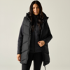 Donna con capelli lunghi scuri che indossa un lungo gilet imbottito nero con cappuccio sopra una giacca nera con cerniera e leggings neri, in piedi su sfondo chiaro