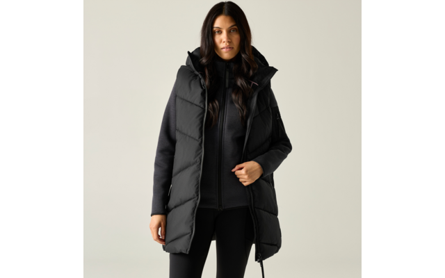 Donna con capelli lunghi scuri che indossa un lungo gilet imbottito nero con cappuccio sopra una giacca nera con cerniera e leggings neri, in piedi su sfondo chiaro