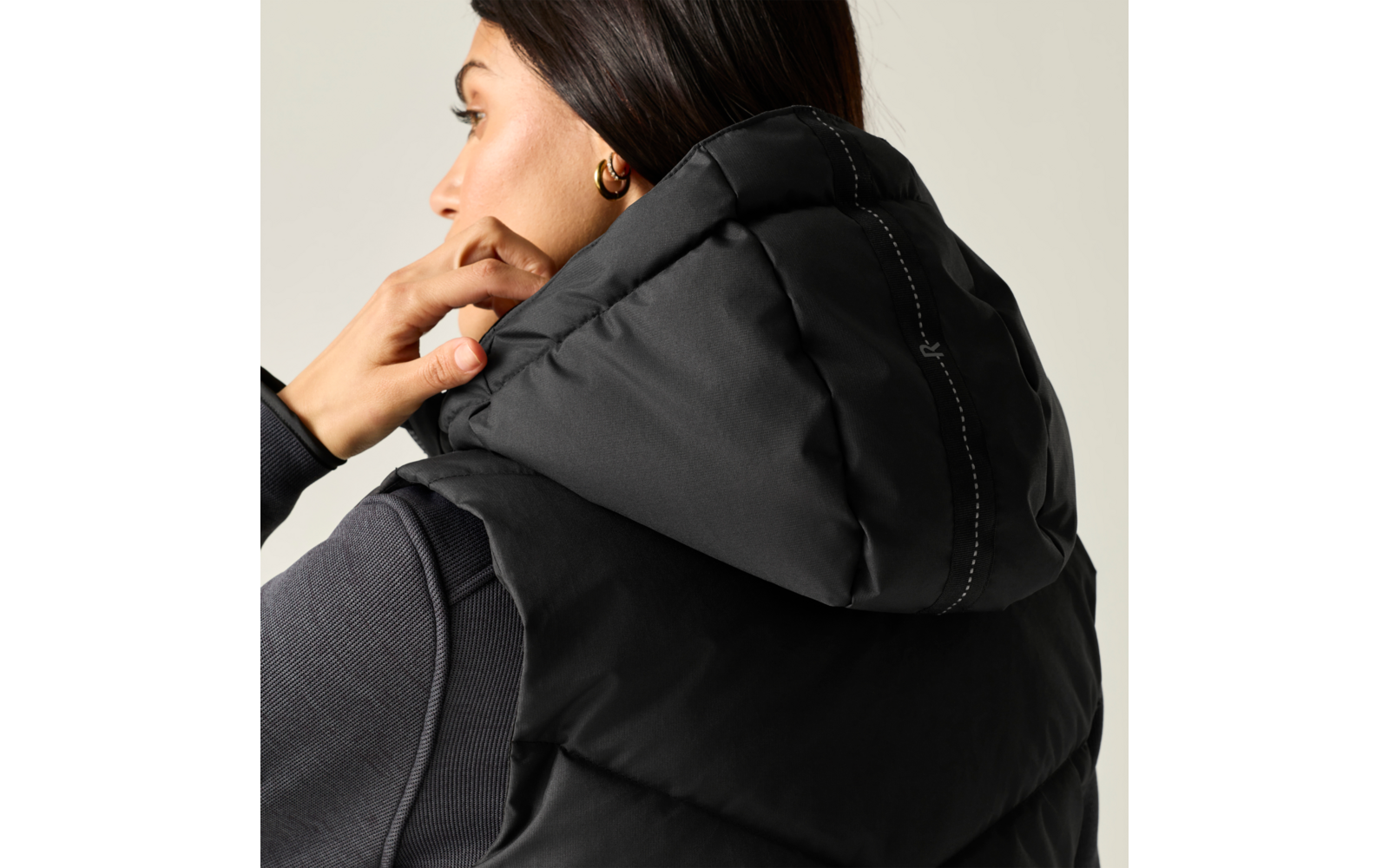 Persona di profilo con capelli scuri raccolti, indossa un gilet imbottito nero con cappuccio e una maglia a maniche lunghe scura, mano sul cappuccio, orecchino a cerchio dorato, sfondo chiaro neutro