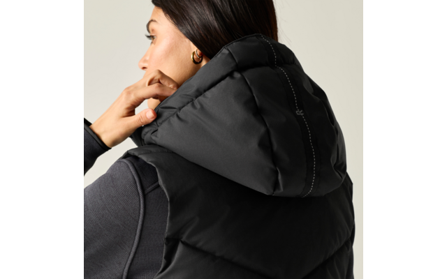 Persona di profilo con capelli scuri raccolti, indossa un gilet imbottito nero con cappuccio e una maglia a maniche lunghe scura, mano sul cappuccio, orecchino a cerchio dorato, sfondo chiaro neutro
