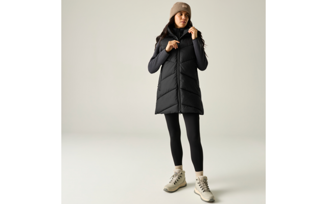 Persona in piedi in uno studio chiaro, indossa un gilet imbottito senza maniche nero lungo, una maglia nera a maniche lunghe, leggings neri, un berretto beige e scarpe da trekking beige; mani vicino al colletto
