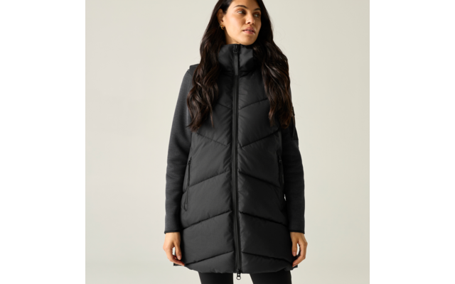 Persona con gilet imbottito lungo nero con collo alto e cerniera frontale, maglia a maniche lunghe scura, capelli lunghi e scuri, in piedi di fronte a uno sfondo chiaro uniforme, mani lungo i fianchi