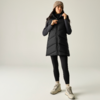 Persona in piedi in uno studio chiaro, indossa un gilet imbottito senza maniche nero lungo, una maglia nera a maniche lunghe, leggings neri, un berretto beige e scarpe da trekking beige; mani vicino al colletto