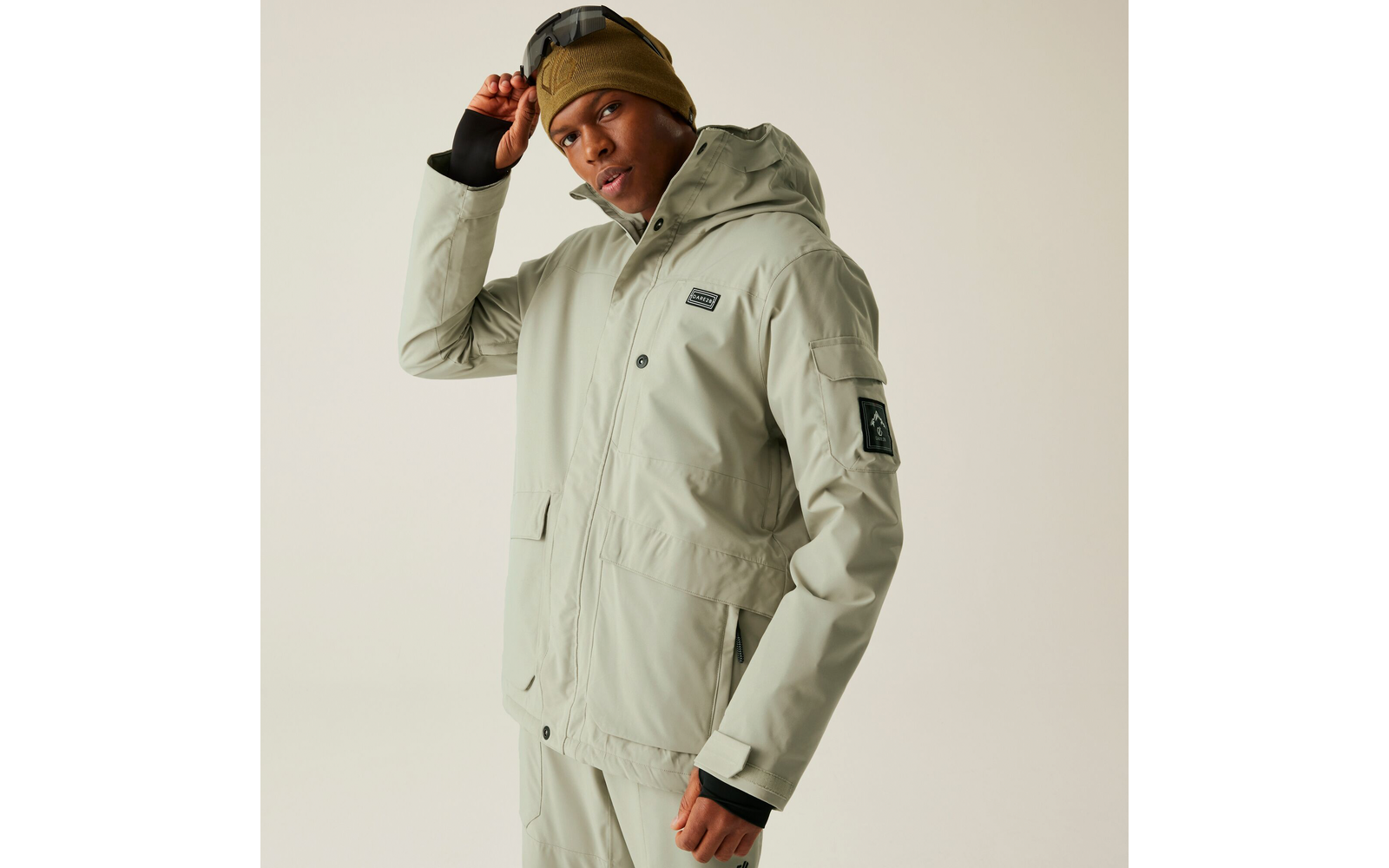 Person in hellgrauem Outdoor-Outfit mit beiger Mütze vor grauem Hintergrund