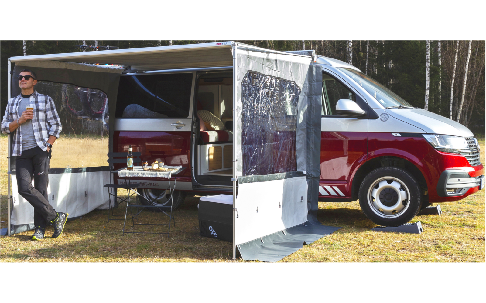 Roter Campingbus mit offener Markise und Anbau, Mann steht daneben auf Wiese vor Wald