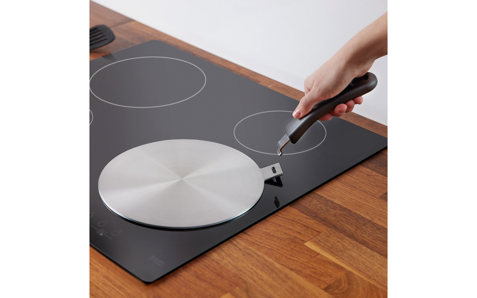 Main tenant une poignée noire au-dessus d'un fond de poêle rond en acier inoxydable posé sur une plaque vitrocéramique noire avec zones de cuisson circulaires visibles et plan de travail en bois