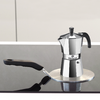 Cafetière moka/expresso argentée sur une plaque métallique ronde sur une plaque de cuisson en verre noir, poignée noire à gauche et reflets, fond clair