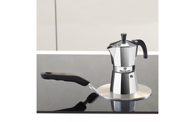 Cafetière moka/expresso argentée sur une plaque métallique ronde sur une plaque de cuisson en verre noir, poignée noire à gauche et reflets, fond clair