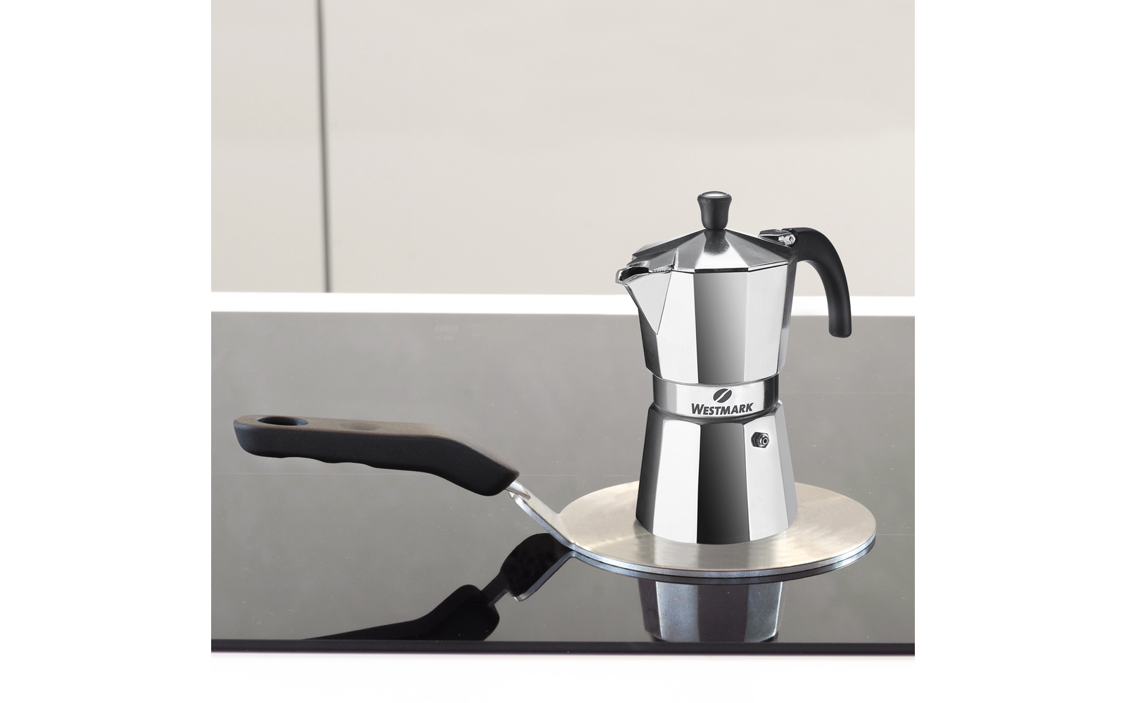 Cafetière moka/expresso argentée sur une plaque métallique ronde sur une plaque de cuisson en verre noir, poignée noire à gauche et reflets, fond clair