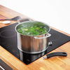 Casserole en acier inoxydable avec eau bouillante et morceaux de légumes verts sur une plaque à induction noire ; poignée amovible et ustensiles sur plan de travail en bois