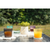 Mesa de madera oscura al aire libre con cuatro vasos acanalados de distintos colores, incluido uno con líquido naranja y uno con líquido marrón oscuro; maceta blanca con planta verde sobre un paño blanco; fondo verde desenfocado