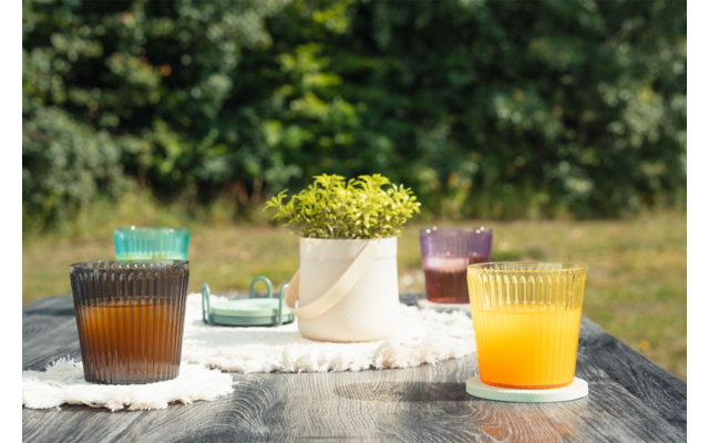 Mesa de madera oscura al aire libre con cuatro vasos acanalados de distintos colores, incluido uno con líquido naranja y uno con líquido marrón oscuro; maceta blanca con planta verde sobre un paño blanco; fondo verde desenfocado