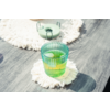 Vaso verde acanalado con líquido verde y cubitos de hielo sobre un posavasos redondo blanco con flecos sobre una superficie de madera gris; en el fondo otro posavasos y un objeto redondo verde