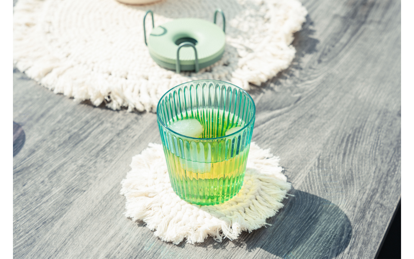 Vaso verde acanalado con líquido verde y cubitos de hielo sobre un posavasos redondo blanco con flecos sobre una superficie de madera gris; en el fondo otro posavasos y un objeto redondo verde