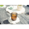 Vaso marrón acanalado con líquido naranja sobre posavasos blanco con flecos en mesa de madera gris; al fondo maceta, objeto verde y otro vaso