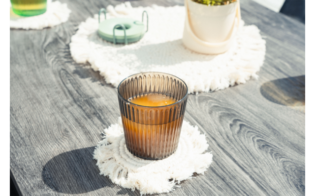 Vaso marrón acanalado con líquido naranja sobre posavasos blanco con flecos en mesa de madera gris; al fondo maceta, objeto verde y otro vaso
