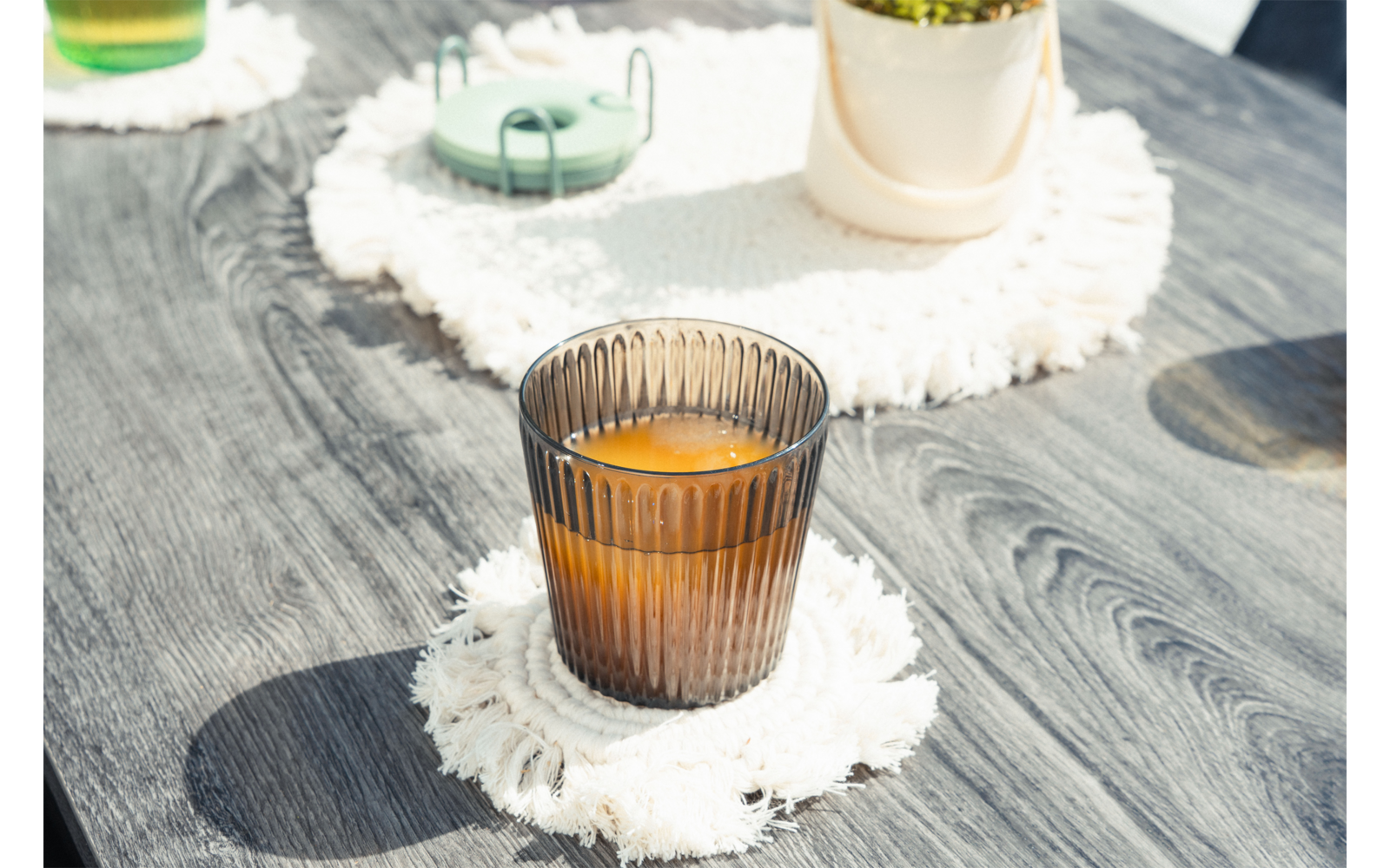 Vaso marrón acanalado con líquido naranja sobre posavasos blanco con flecos en mesa de madera gris; al fondo maceta, objeto verde y otro vaso