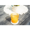 Vaso acanalado con líquido naranja sobre un posavasos azul claro en una mesa de madera gris; tapete blanco de ganchillo y maceta blanca en el fondo