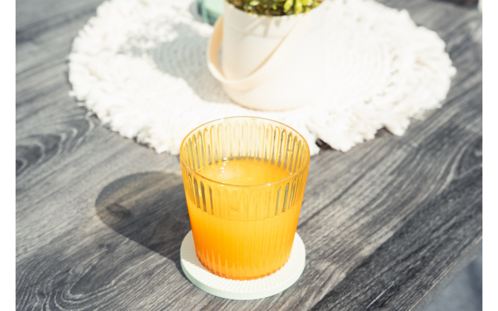 Vaso acanalado con líquido naranja sobre un posavasos azul claro en una mesa de madera gris; tapete blanco de ganchillo y maceta blanca en el fondo