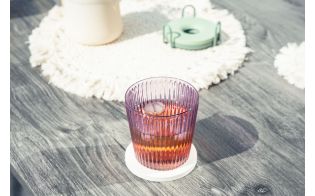 Vaso morado acanalado con líquido ámbar y cubitos de hielo sobre un posavasos blanco en una superficie de madera gris; al fondo un salvamantel redondo blanco texturizado, un recipiente claro y un objeto redondo verde
