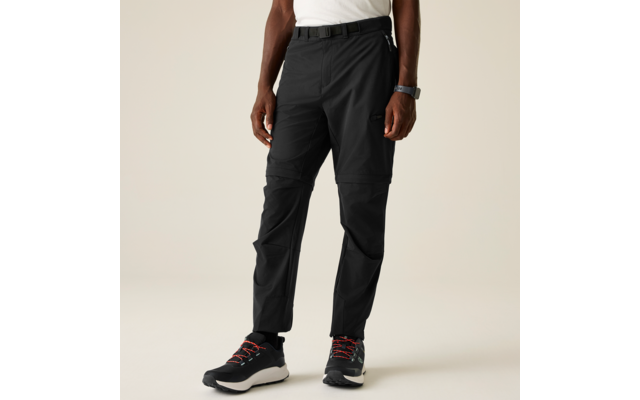 Parte inferior del cuerpo de una persona sobre fondo beige, camiseta blanca, cinturón negro, pantalones negros con costura visible y cremallera a la altura de las rodillas y bolsillo lateral en el muslo, reloj en la muñeca izquierda, zapatillas deportivas negras con suela blanca y cordones rojos