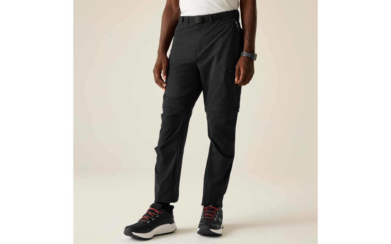 Parte inferior del cuerpo de una persona sobre fondo beige, camiseta blanca, cinturón negro, pantalones negros con costura visible y cremallera a la altura de las rodillas y bolsillo lateral en el muslo, reloj en la muñeca izquierda, zapatillas deportivas negras con suela blanca y cordones rojos